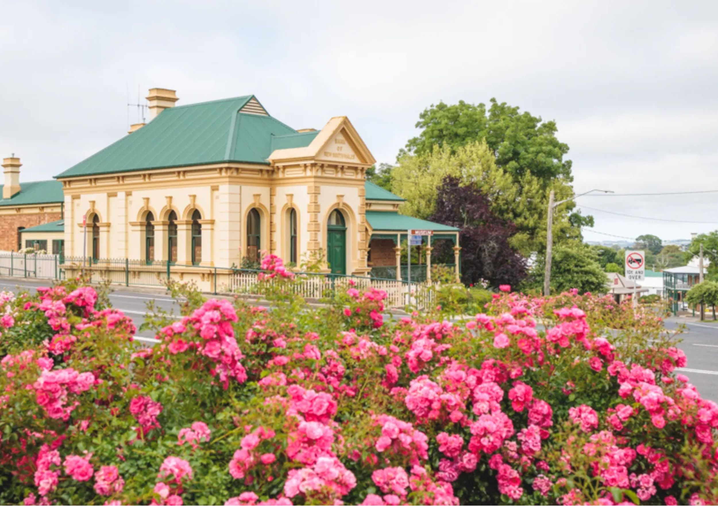 Historic Rosebank Guest House in Millthorpe, NSW, featuring a heritage-style building with a green roof and arched windows, framed by vibrant blooming rose gardens along the roadside.