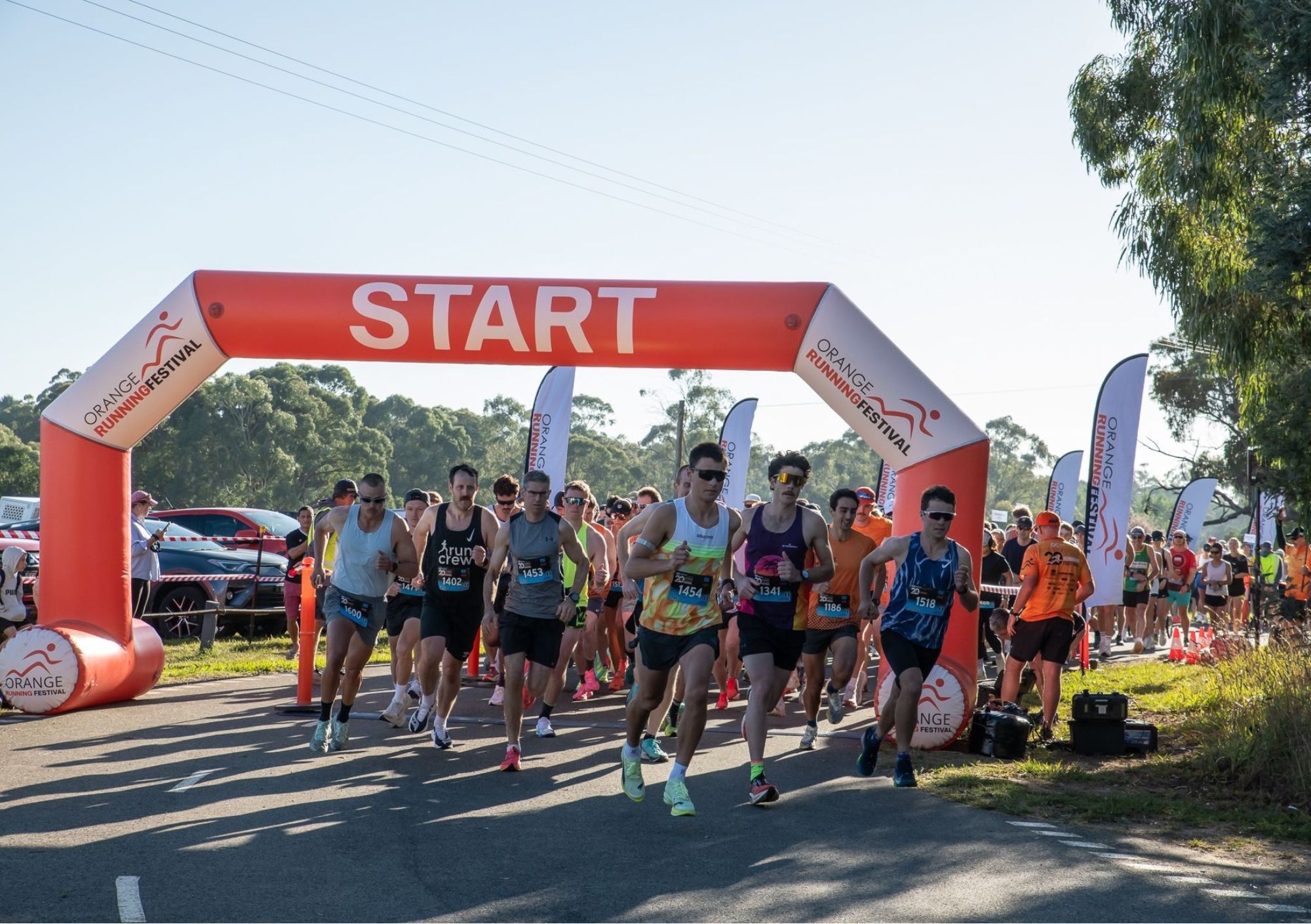 A group of runners begins a race at the Orange Running Festival, passing under a large red “START” inflatable arch on a sunny day, with spectators, flags, and trees in the background.